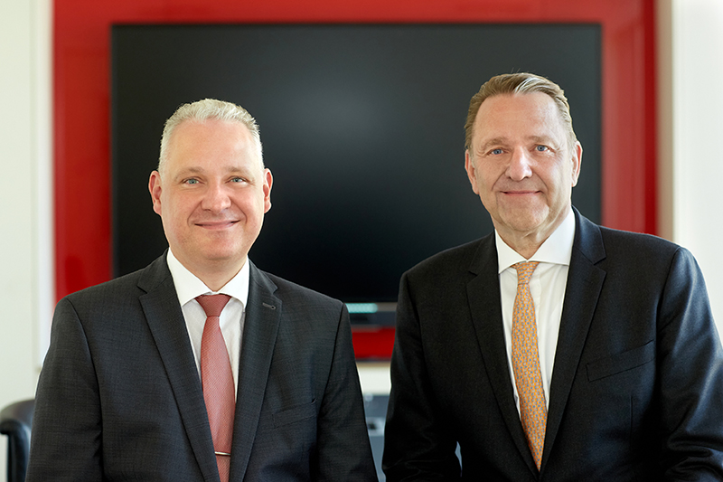 Zwei Männer im Anzug sitzend, lächeln in die Kamera, weiß/roter Hintergrund