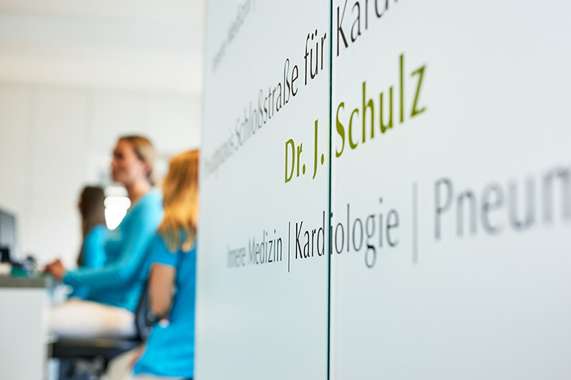 Zwei Mitarbeiterinnen im blauen Kittel sind klein und unscherf im Hintergrund zu sehen. Vorne der Schriftzug: Praxis Dr. J. Schulz